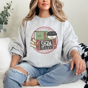 May include: A gray sweatshirt with a graphic design of a stack of books, a coffee cup, and a video game console. The graphic is surrounded by a pink circle with a dashed line border. The text "Stay Cozy" is written in black on a white background.