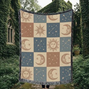 May include: A woven throw blanket featuring a celestial design with suns, moons, stars, and constellations in a patchwork pattern. The color palette includes beige, brown, and blue squares, with a fringed edge.