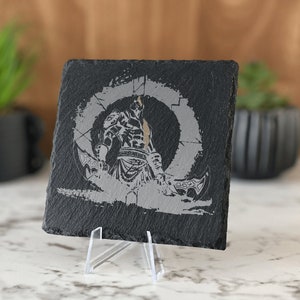 May include: A black slate coaster with a silver-colored image of a warrior holding a weapon. The warrior is standing in a circle with a symbol in the center. The coaster is on a white stand.