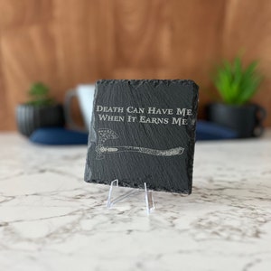 May include: Black slate coaster with a white engraved axe and the text "Death can have me when it earns me".