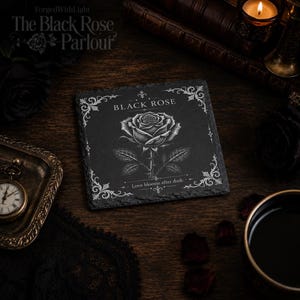 Pu&ograve; includere: Un sottobicchiere quadrato in ardesia nera con un'illustrazione incisa in bianco di una singola rosa al centro. Il testo "BLACK ROSE" appare sopra il fiore e "Love blooms after dusk." &egrave; stampato sotto, incorniciato da filigrane bianche agli angoli.