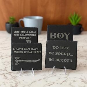 Puede incluir: Cuatro posavasos de pizarra negra con texto grabado. Uno dice "¿ERES UNA PERSONA TRANQUILA Y RAZONABLE?", otro dice "BOY", un tercero dice "LA MUERTE PUEDE TENER CUANDO SE LO GANA" y el último dice "NO TE ARREPIENTAS... SÉ MEJOR".