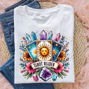 Può includere: Maglietta bianca con un'illustrazione colorata di carte dei tarocchi, cristalli e fiori. Il testo "Tarot Reader" è stampato sulla maglietta.