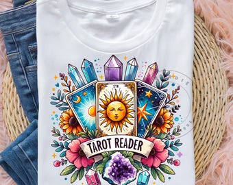 May include: White t-shirt featuring a colourful illustration of tarot cards, crystals, and flowers. The text "Tarot Reader" is printed on the shirt.