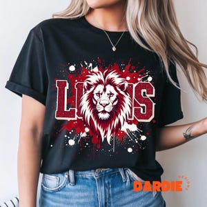 May include: Black t-shirt featuring a lion's head graphic with the word "LIONS" in red and white lettering. The design includes red and white paint splatter effects, creating a bold and dynamic look. The shirt is a classic crew neck.