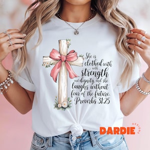 May include: White t-shirt with a pink ribbon tied around a wooden cross. The text on the shirt reads "She is clothed with strength and dignity, and she laughs without fear of the future. Proverbs 31:25".