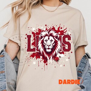 May include: Beige t-shirt featuring a red and white lion graphic with the word "LIONS" in a bold, collegiate font. The design includes a splatter effect in red, adding a dynamic visual element. The shirt is a casual style.