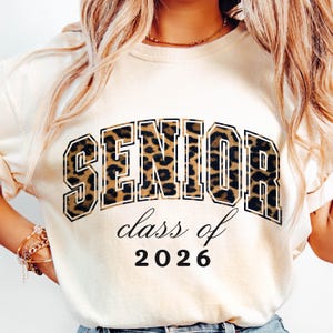 May include: Cream-colored sweatshirt with the word "SENIOR" in large, leopard print letters. Below, the text "class of 2026" is printed in a cursive font, with the year in a bold, block font. A stylish top for the graduating class of 2026.