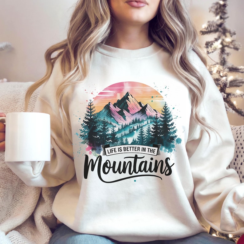Op de afbeelding: Een wit sweatshirt met een aquarelontwerp van een bergketen met een zonsondergang op de achtergrond. De tekst "Life is better in the Mountains" is onder het ontwerp gedrukt.