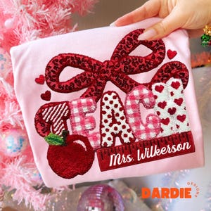 May include: A light pink t-shirt with a red bow and the word "TEACH" embroidered in various patterns. A red apple and the name "Mrs. Wilkerson" are also embroidered on the shirt. The shirt is a teacher-themed item.