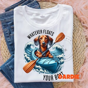 May include: White t-shirt featuring a dachshund in a blue kayak paddling through waves. The text "Whatever floats your boat" is above the kayak and the text "DARDIE" is below the kayak.
