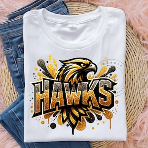May include: White t-shirt with a gold glitter design of a hawk with the word "HAWKS" in gold glitter letters.