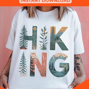 Op de afbeelding: Wit T-shirt met een grafisch ontwerp van het woord "HIKING" in kleurrijke letters. Elke letter is gevuld met een natuursc&egrave;ne, waaronder bomen, bergen en een meer. De tekst "DARDIE DESIGNS" staat onderaan het shirt.
