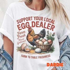 May include: White t-shirt with a red and white graphic design that says "Support Your Local Egg Dealer". The graphic also includes a cartoon illustration of a hen with a basket of eggs.