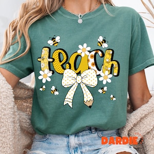 May include: Teal t-shirt with the word "TEACH" in yellow, black, and white, decorated with bees, daisies, and a pencil-shaped bow. The shirt has a relaxed fit and short sleeves.