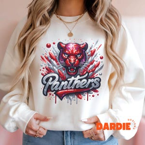May include: White sweatshirt with a red and silver glitter design of a panther with the word "Panthers" in silver glitter. The sweatshirt has a crew neck and long sleeves.