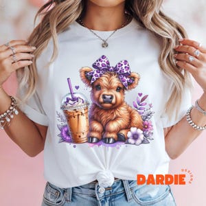 Pu&ograve; includere: Maglietta bianca con un fiocco viola e bianco su una mucca Highland marrone che tiene una tazza di caff&egrave; freddo. Il testo "DARDIE DESIGNS" &egrave; nell'angolo in basso a destra della maglietta.
