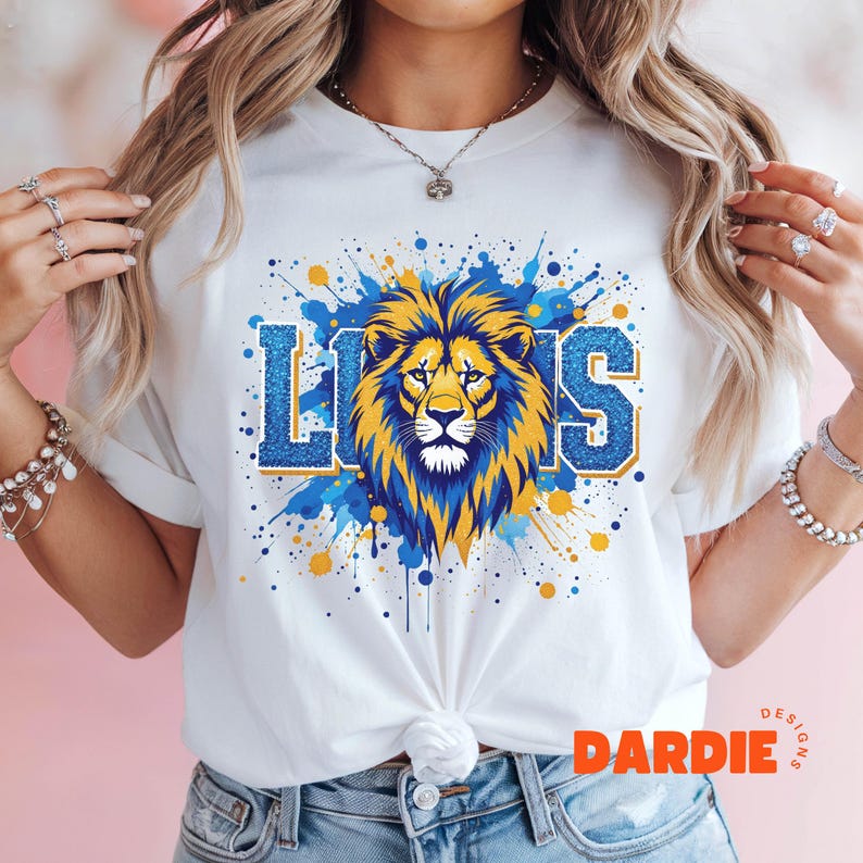 May include: White t-shirt with a blue and yellow lion graphic and the word "LIONS" in blue and yellow glitter. The graphic is surrounded by blue and yellow paint splatters.