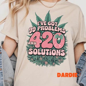May include: Beige t-shirt with a graphic design featuring the text "I've Got 99 Problems And 420 Solutions" in pink and white, surrounded by green cannabis leaves and flowers. The word "DARDIE" is printed in orange.