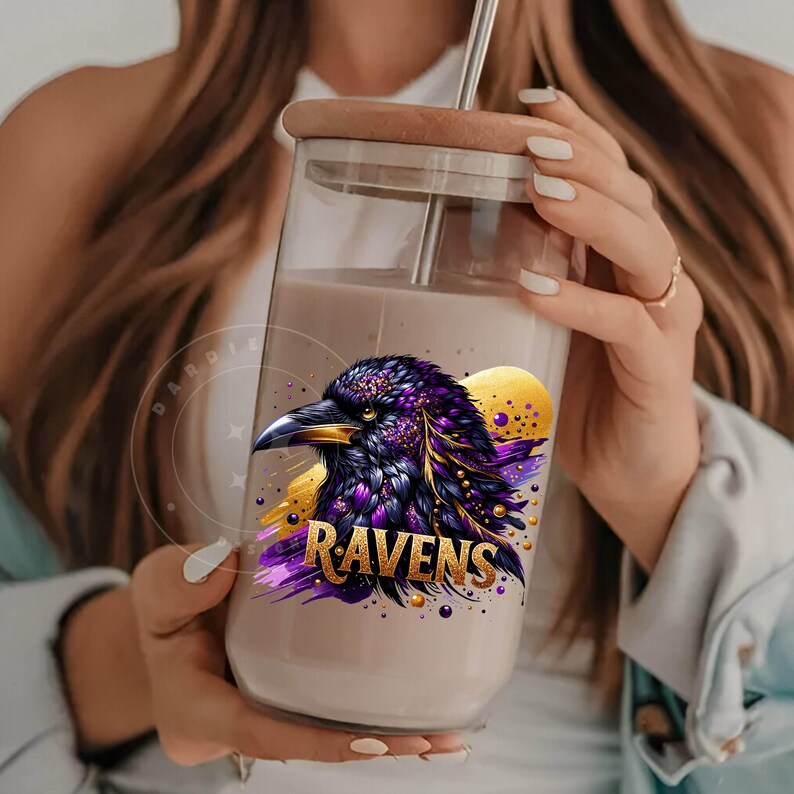 May include: A clear glass tumbler with a wooden lid and a metal straw. The tumbler features a graphic of a purple and black raven with gold accents and the word "RAVENS" in gold lettering. The tumbler is filled with a light-colored liquid.