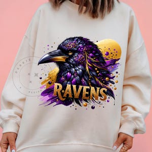 May include: Cream-colored sweatshirt featuring a detailed illustration of a raven with purple and gold accents. The word "RAVENS" is displayed in gold lettering below the bird. The design includes a golden sun-like shape in the background.
