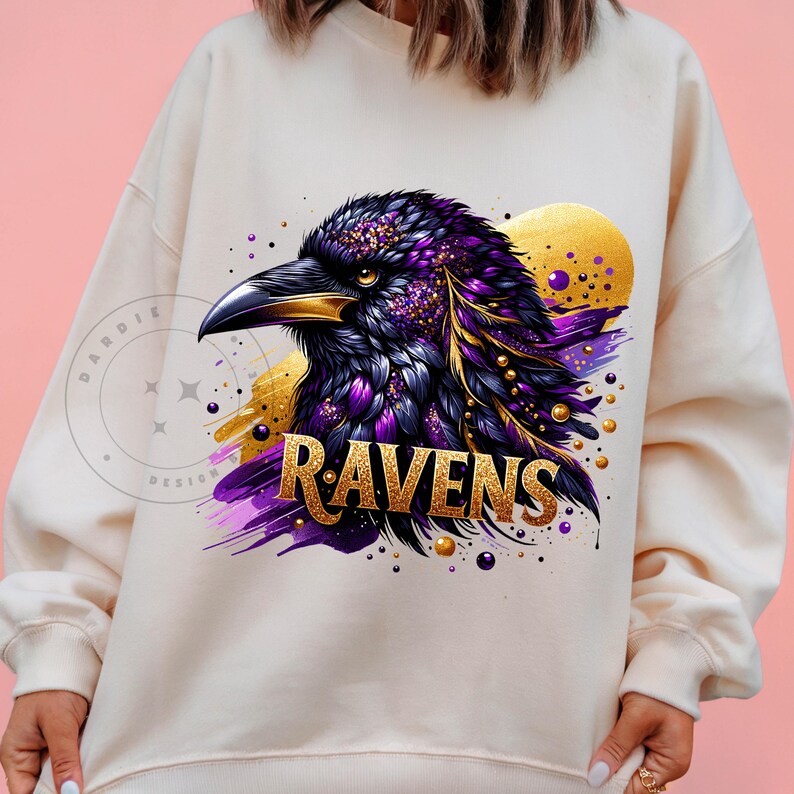 May include: Cream-colored sweatshirt featuring a detailed illustration of a raven with purple and gold accents. The word "RAVENS" is displayed in gold lettering below the bird. The design includes a golden sun-like shape in the background.