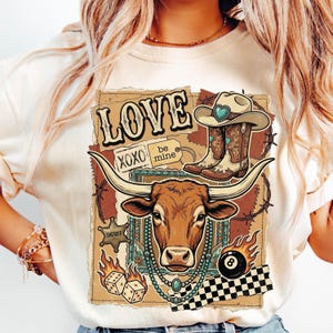 以下が含まれることがあります： クリーム色のTシャツ。ウエスタンをテーマにしたグラフィックデザイン。ロングホーン、カウボーイブーツ、カウボーイハット、サイコロ、そして「LOVE」の文字が描かれています。「XOXO」と「be mine」の文字も含まれています。