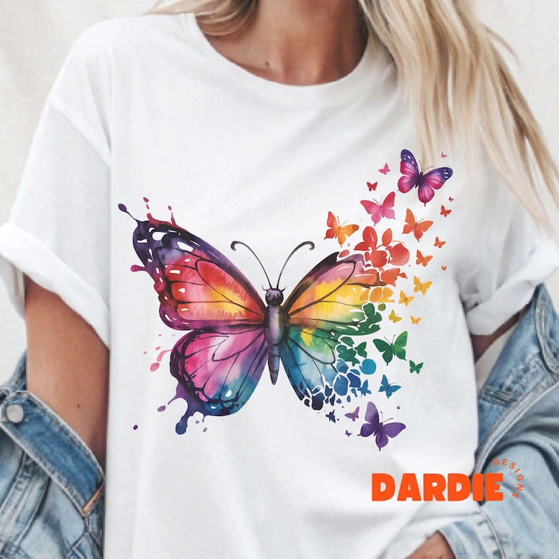 May include: A white t-shirt with a colorful watercolor butterfly design. The butterfly is made up of many different colors, including red, orange, yellow, green, blue, and purple. The butterfly is surrounded by smaller butterflies, which are also made up of different colors. The shirt has the word "DARDIE" printed on the bottom right corner.
