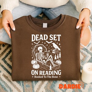 May include: A brown sweatshirt with a white graphic that reads "DEAD SET ON READING" with a skeleton in a coffin, pumpkins, bats, a raven, and candles. The text "Booked To The Bone" is also included.