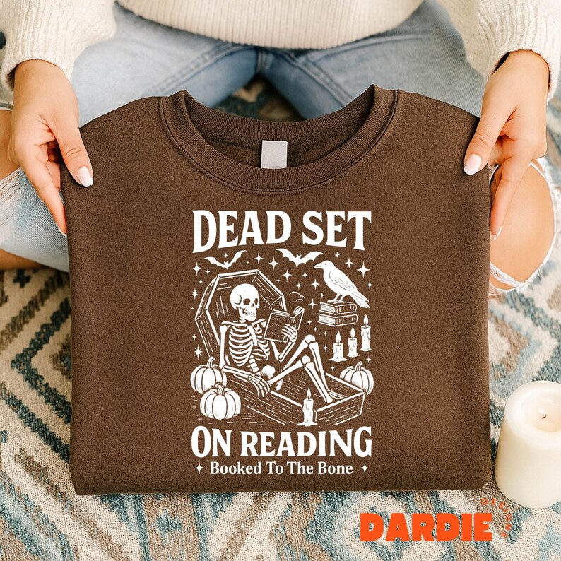 May include: A brown sweatshirt with a white graphic that reads "DEAD SET ON READING" with a skeleton in a coffin, pumpkins, bats, a raven, and candles. The text "Booked To The Bone" is also included.