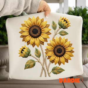 Puede incluir: Cojín color crema con un diseño bordado detallado de girasoles. Los girasoles tienen pétalos amarillos, centros marrones y hojas verdes. Los tallos están hechos de un material similar a una cuerda, creando un efecto tridimensional.