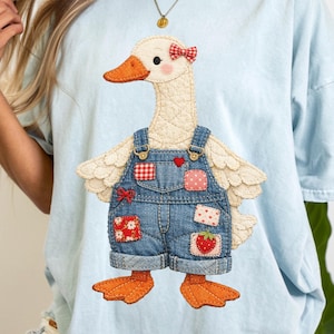 May include: Light blue t-shirt featuring a whimsical goose design. The goose is wearing denim overalls with patches and a red bow. The goose has an orange beak and feet, and a quilted body.