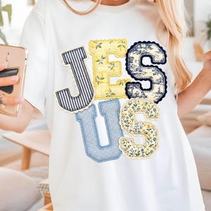Può includere: T-shirt bianca con la scritta "JESUS" in lettere applicate colorate. La "J" è a righe, la "E" ha limoni, la "S" presenta un motivo toile de Jouy blu e bianco, la "U" ha pois blu e l'ultima "S" ha un motivo floreale.