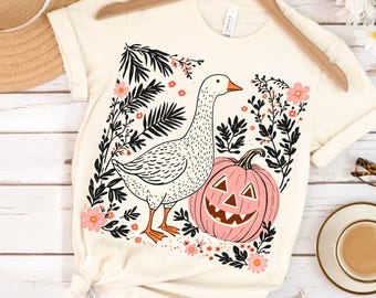 May include: Cream-colored t-shirt featuring a white goose with orange feet and beak, standing next to a pink jack-o'-lantern. The design includes black floral elements and pink flowers, creating a fall or Halloween theme.