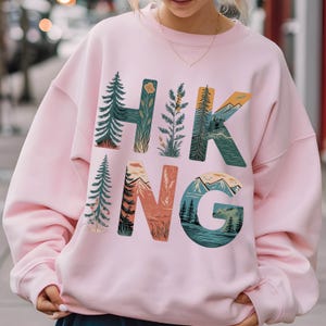Op de afbeelding: Roze sweatshirt met een grafisch ontwerp van het woord "HIKING" met natuurtaferelen die in de letters zijn verwerkt.