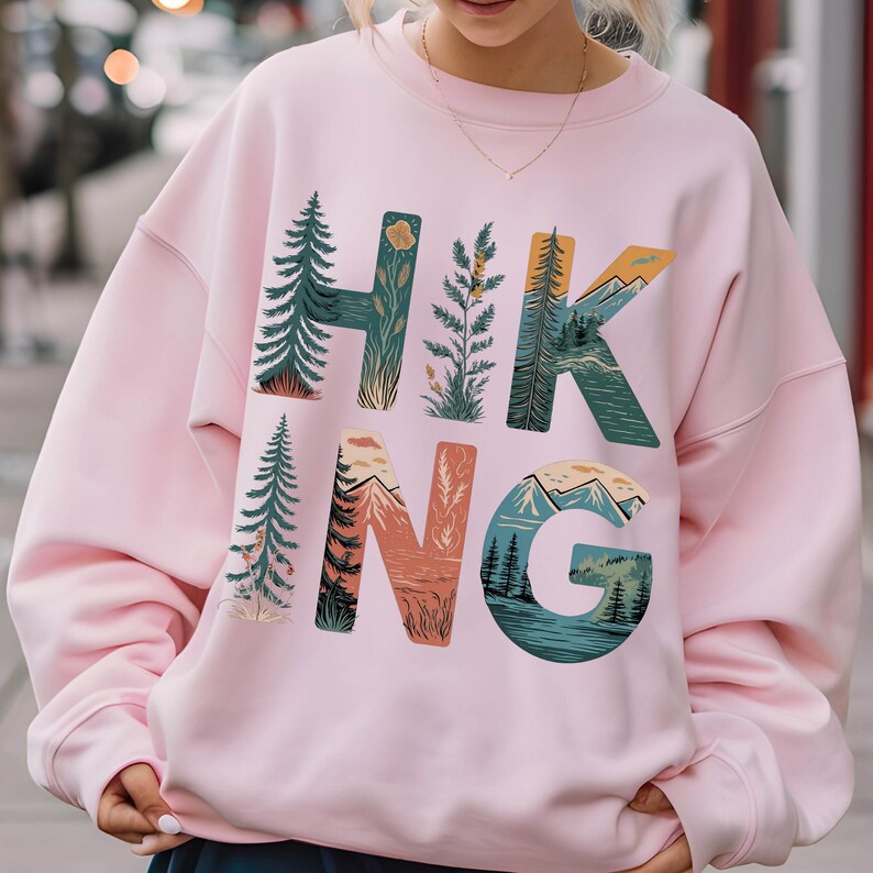 Op de afbeelding: Roze sweatshirt met een grafisch ontwerp van het woord "HIKING" met natuurtaferelen die in de letters zijn verwerkt.