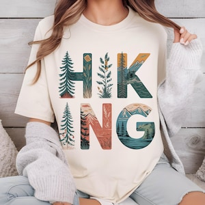 Op de afbeelding: Een cr&egrave;mekleurig T-shirt met het woord "HIKING" in grote letters bedrukt. Elke letter is gevuld met een natuursc&egrave;ne, waaronder bomen, bergen en een meer.