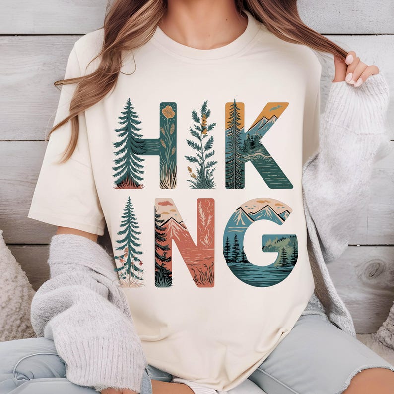 Op de afbeelding: Een cr&egrave;mekleurig T-shirt met het woord "HIKING" in grote letters bedrukt. Elke letter is gevuld met een natuursc&egrave;ne, waaronder bomen, bergen en een meer.