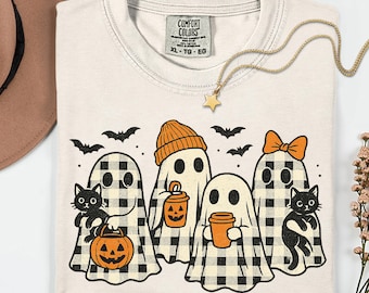 May include: Cream-colored t-shirt featuring a Halloween-themed graphic. The design includes ghosts in buffalo plaid, black cats, bats, and jack-o'-lanterns. A gold star necklace and a Comfort Colors tag are also visible.
