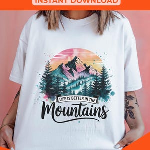 Op de afbeelding: Een wit T-shirt met een aquarelontwerp van bergen, bomen en een zonsondergang. De tekst "Life is better in the Mountains" is geschreven in zwarte cursieve letters.