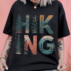 Op de afbeelding: Zwart T-shirt met een grafisch ontwerp van het woord "HIKING" met natuurtaferelen die in elke letter zijn verwerkt.