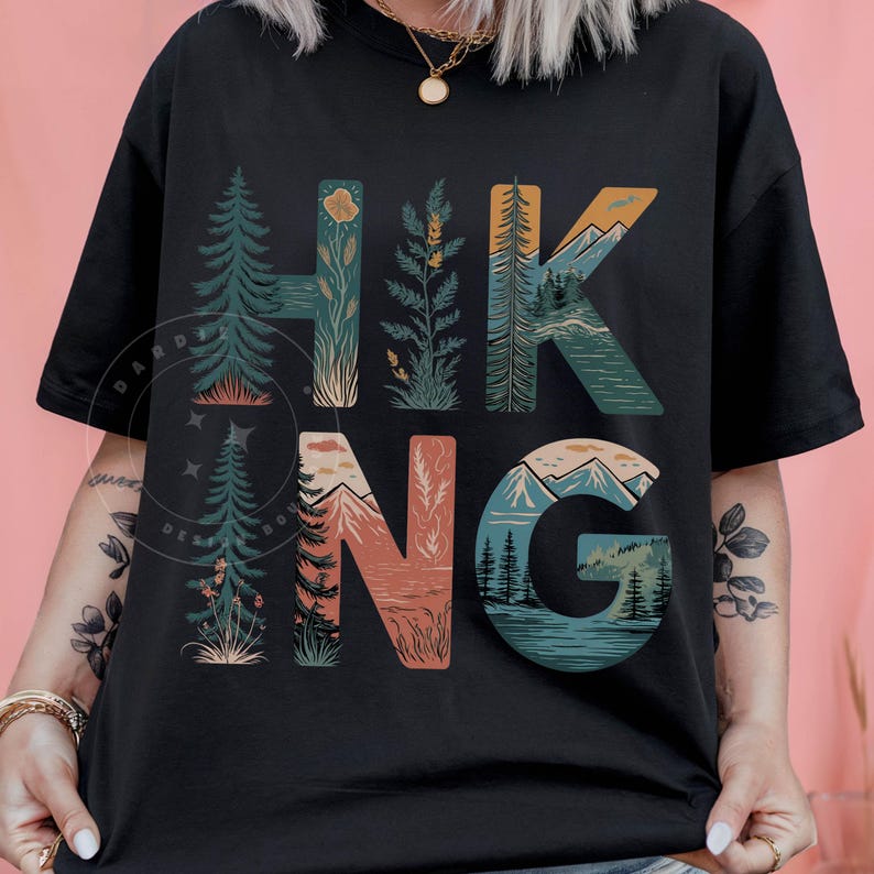 Op de afbeelding: Zwart T-shirt met een grafisch ontwerp van het woord "HIKING" met natuurtaferelen die in elke letter zijn verwerkt.