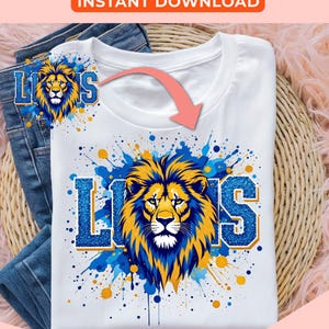 May include: A white t-shirt with a blue and gold lion graphic and the word "Lions" in blue and gold glitter. The graphic is surrounded by blue and gold paint splatters.