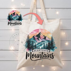 Op de afbeelding: Een witte canvas tote bag met een aquarelontwerp van bergen, bomen en een zonsondergang. De tekst "Life is better in the Mountains" is in zwart op de tas gedrukt.