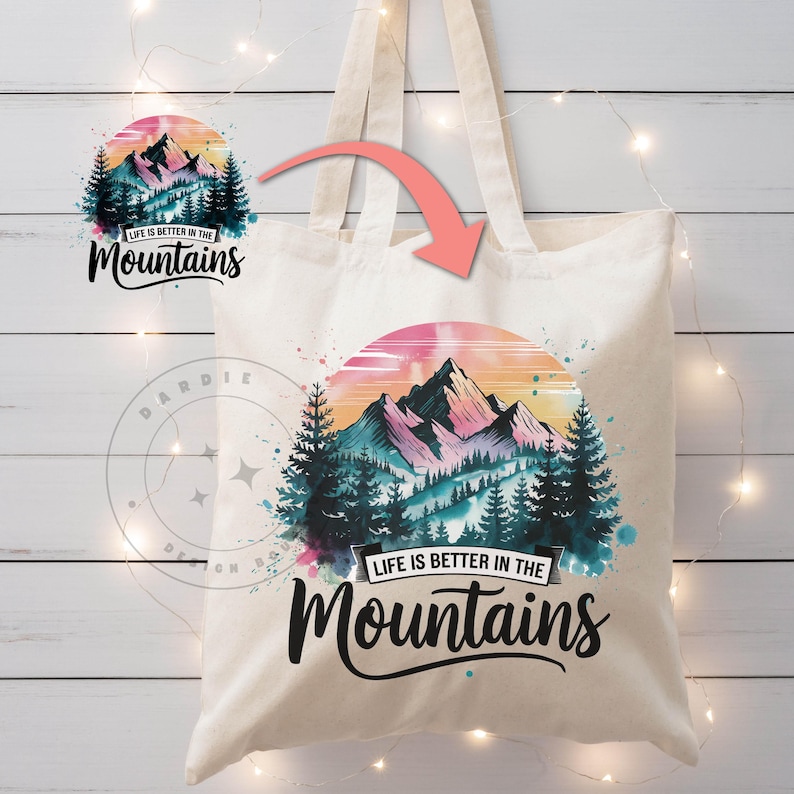 Op de afbeelding: Een witte canvas tote bag met een aquarelontwerp van bergen, bomen en een zonsondergang. De tekst "Life is better in the Mountains" is in zwart op de tas gedrukt.