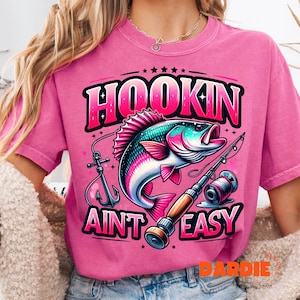 May include: Pink t-shirt with a graphic design featuring a colorful fish, fishing rod, and the text "Hookin Ain't Easy". The design includes an anchor and stars. The shirt is a solid pink color.