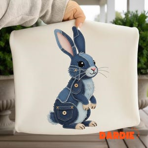 Può includere: Un cuscino color crema con un design a coniglio patchwork in denim. Il coniglio ha toppe in denim blu, dettagli di bottoni e un naso e orecchie interne rosa. Il cuscino è tenuto da una persona.