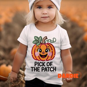 Puede incluir: Una camiseta blanca con un diseño de calabaza sonriente, una hoja verde y rizos. El texto "PICK OF THE PATCH" está impreso debajo. La camiseta la lleva un niño pequeño.