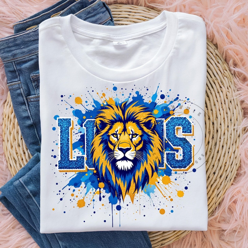 May include: White t-shirt with a blue and gold lion graphic and the word "LIONS" in blue glitter letters. The graphic is surrounded by blue and gold paint splatters.