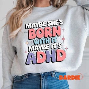 May include: A light grey sweatshirt featuring the text "MAYBE SHE'S BORN WITH IT MAYBE IT'S ADHD" in a colourful, playful font. The design includes pink, blue, and purple lettering with star accents. The sweatshirt is a casual style.